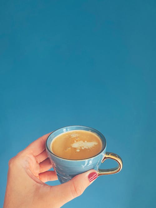 Female hand with red manicure demonstrating cup of fresh cappuccino with froth on blue background