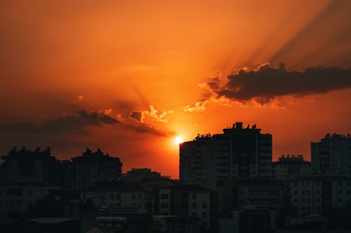 Free Silhouette of city buildings against a stunning orange sunset sky with sunrays breaking through clouds. Stock Photo