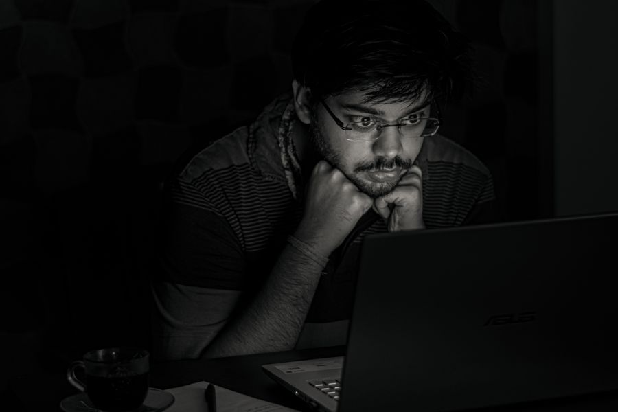 A bearded man intensely focuses on his laptop screen with hands on chin, in a dimly lit room, representing late-night work.