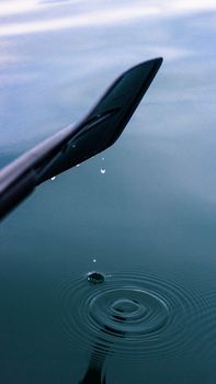 Close-up of paddle dripping water droplets into serene lake creating ripples.