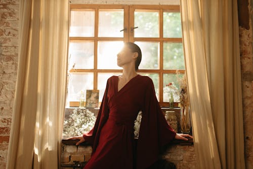 Woman in Red Long Sleeves Dress Leaning on a Windowsill while Posing at the Camera