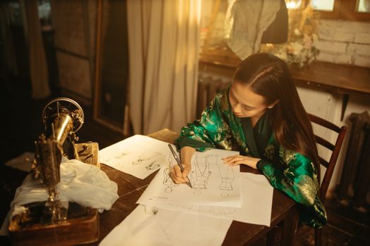 An adult woman sketching fashion designs at a vintage sewing table, indoors.