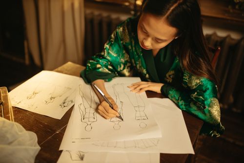 Free Asian woman fashion designer sketching clothing designs at desk, showcasing creativity and concentration. Stock Photo