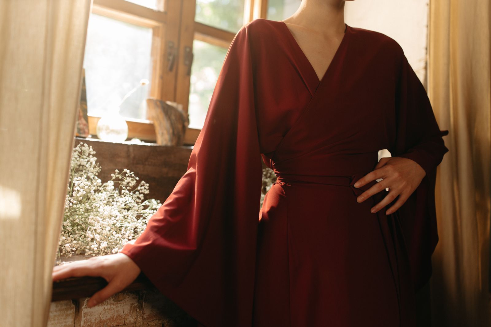 Woman in a belted burgundy wrap dress stands by a sunlit window with white flowers