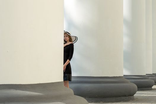 A stylish young woman in a black dress and sun hat stands beside white columns, creating a striking contrast.