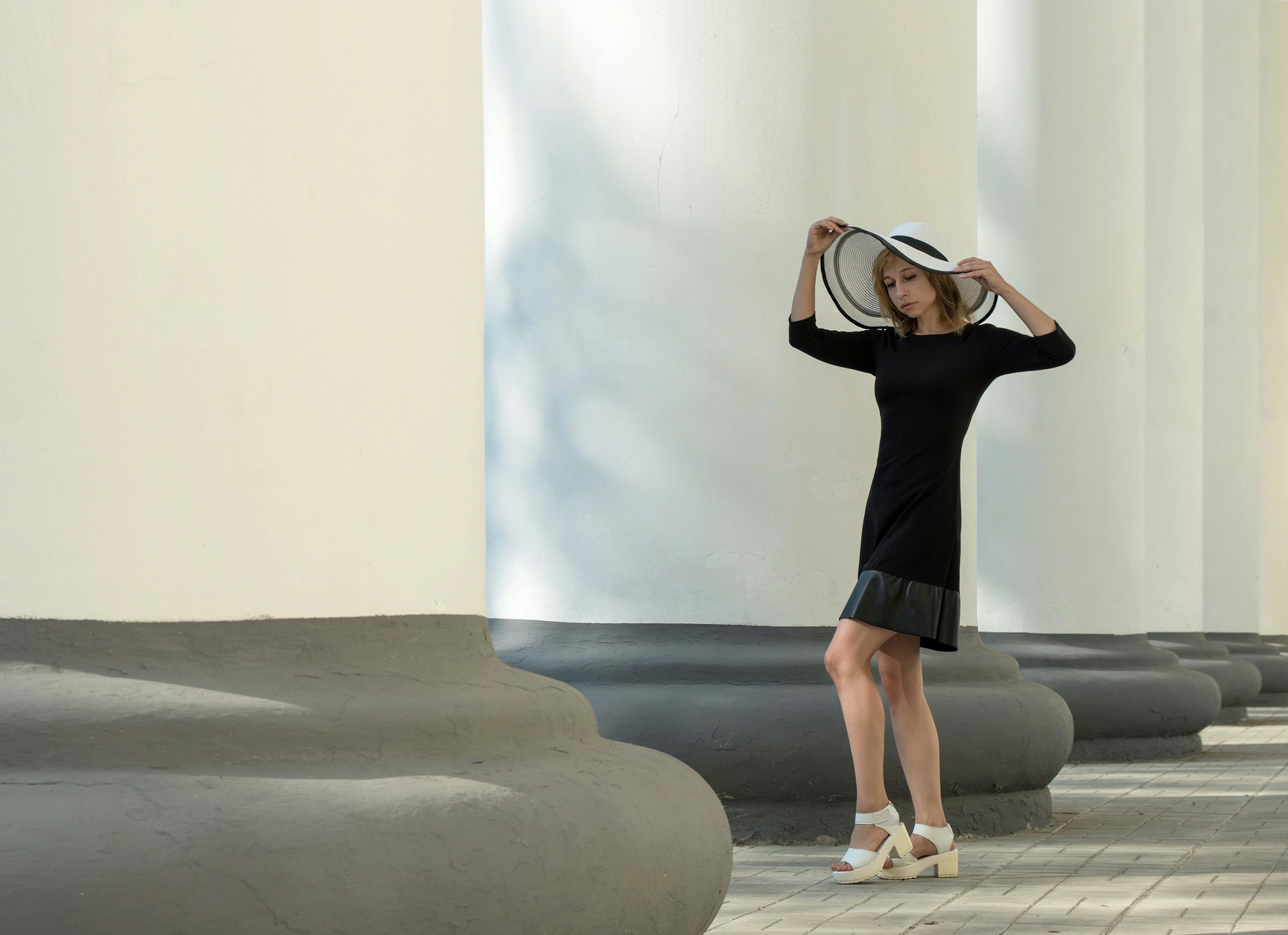A woman in a black dress and hat poses elegantly by grand pillars outdoors.