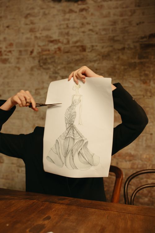 Free A designer showcasing a fashion sketch while holding scissors in a creative workspace. Stock Photo