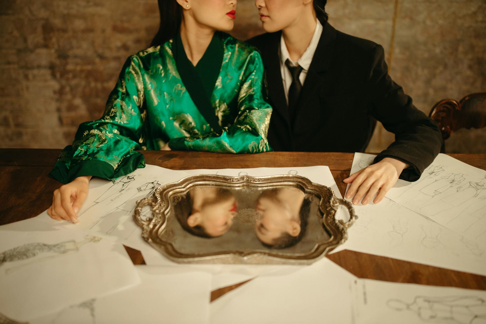 Two fashion designers in elegant attire with reflections in a mirror, surrounded by sketches.