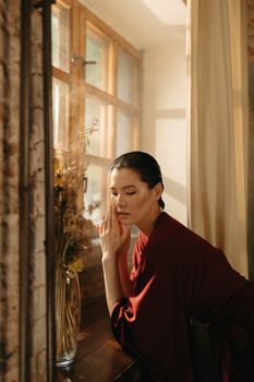A serene woman in a red robe standing by a window, basking in soft natural light.