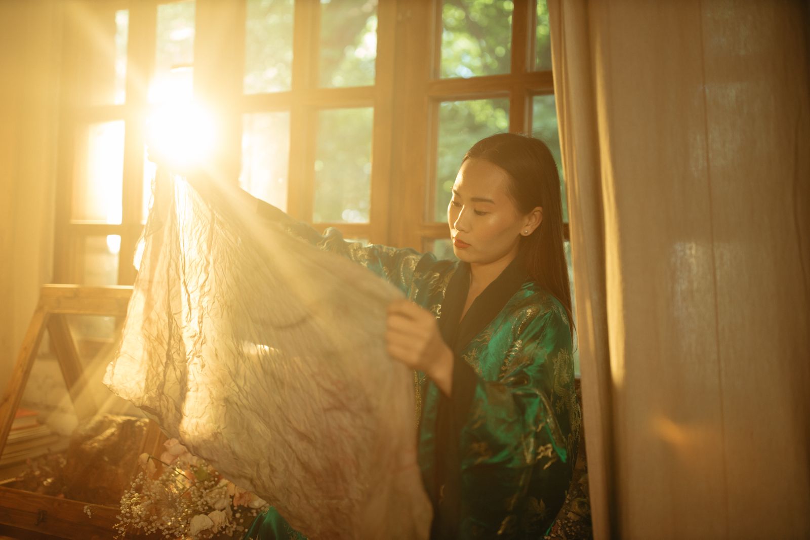 Woman in a green floral robe holding sheer fabric in a sunlit room by tall windows
