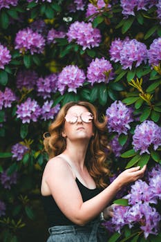 A woman poses among vibrant pink rhododendrons wearing sunglasses, embracing nature's beauty.