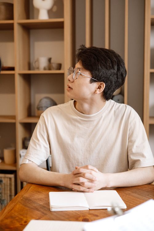 Free A young man wearing glasses sits at a wooden table looking sideways, hands folded. Stock Photo