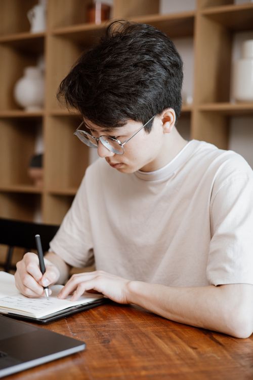 Free Young professional writing with focus at a wooden table in a modern office setting. Stock Photo