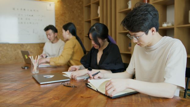 Group of diverse students studying at a wooden table in a modern classroom with laptops and notebooks.