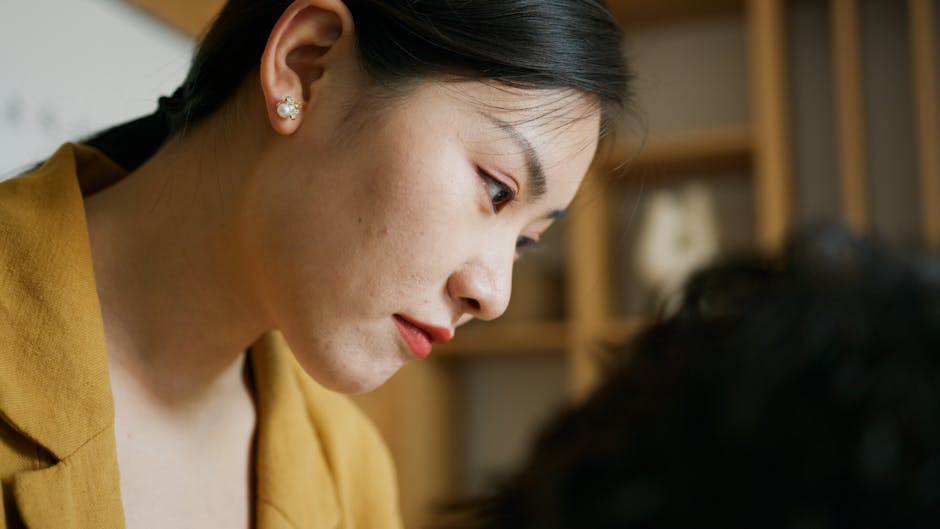 Asian woman in mustard jacket focused intently, indoors close-up.