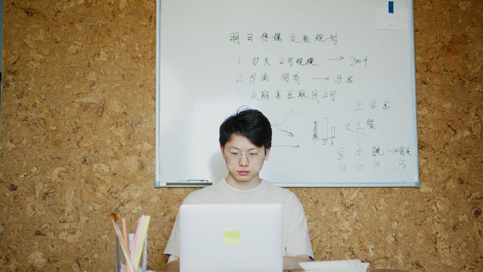 Adult man working on a laptop in a modern office setting with a whiteboard.