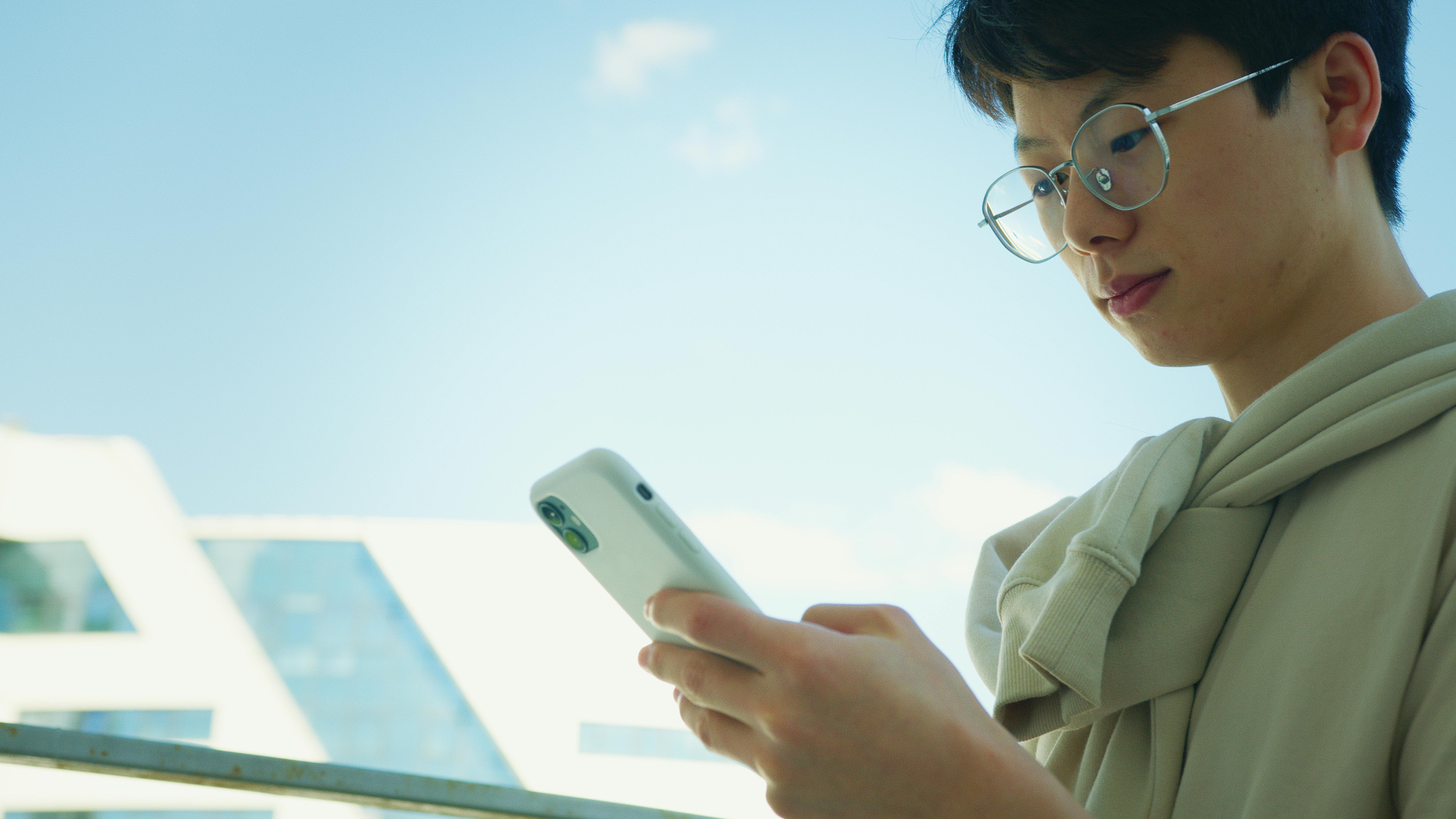 A young adult wearing glasses uses a smartphone outdoors on a sunny day.