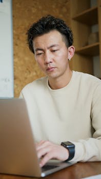 A focused Asian man in a sweater typing on a laptop in a modern indoor office setting.