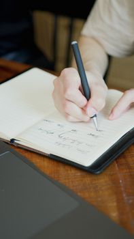 A hand holding a pen writing in an open notebook on a wooden table.