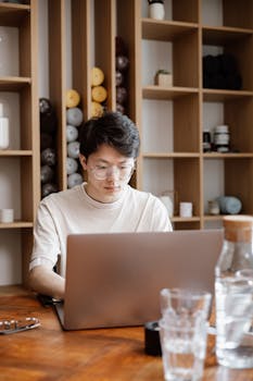 Asian man concentrating on laptop work at a wooden desk with modern shelving in the background.