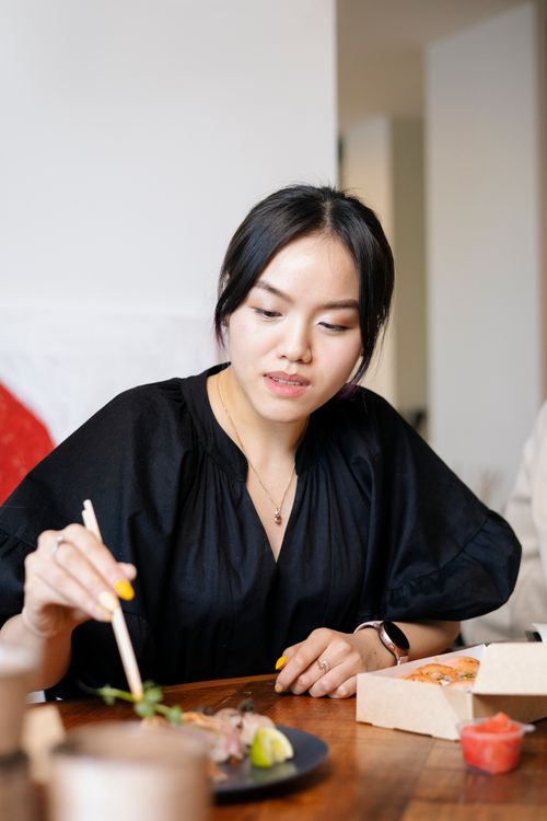 Free Asian woman dining indoors, using chopsticks to enjoy a meal at a wooden table. Stock Photo