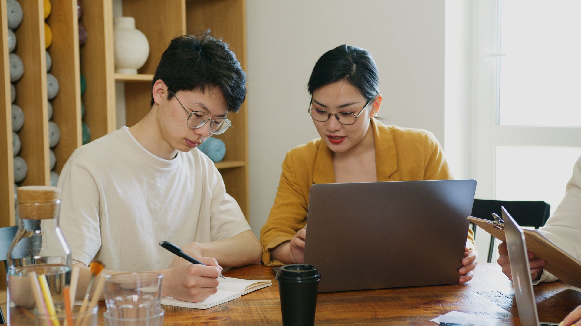 Two professionals working together on a laptop and notebook in a bright office space.