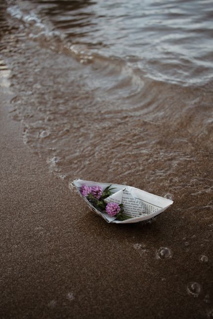 A paper boat holding flowers gently floats on a sandy beach, close to the water's edge.