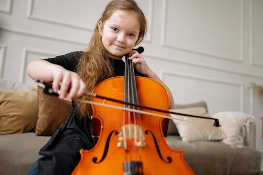 A smiling girl in a black dress plays the cello indoors, showcasing joy and musical talent.