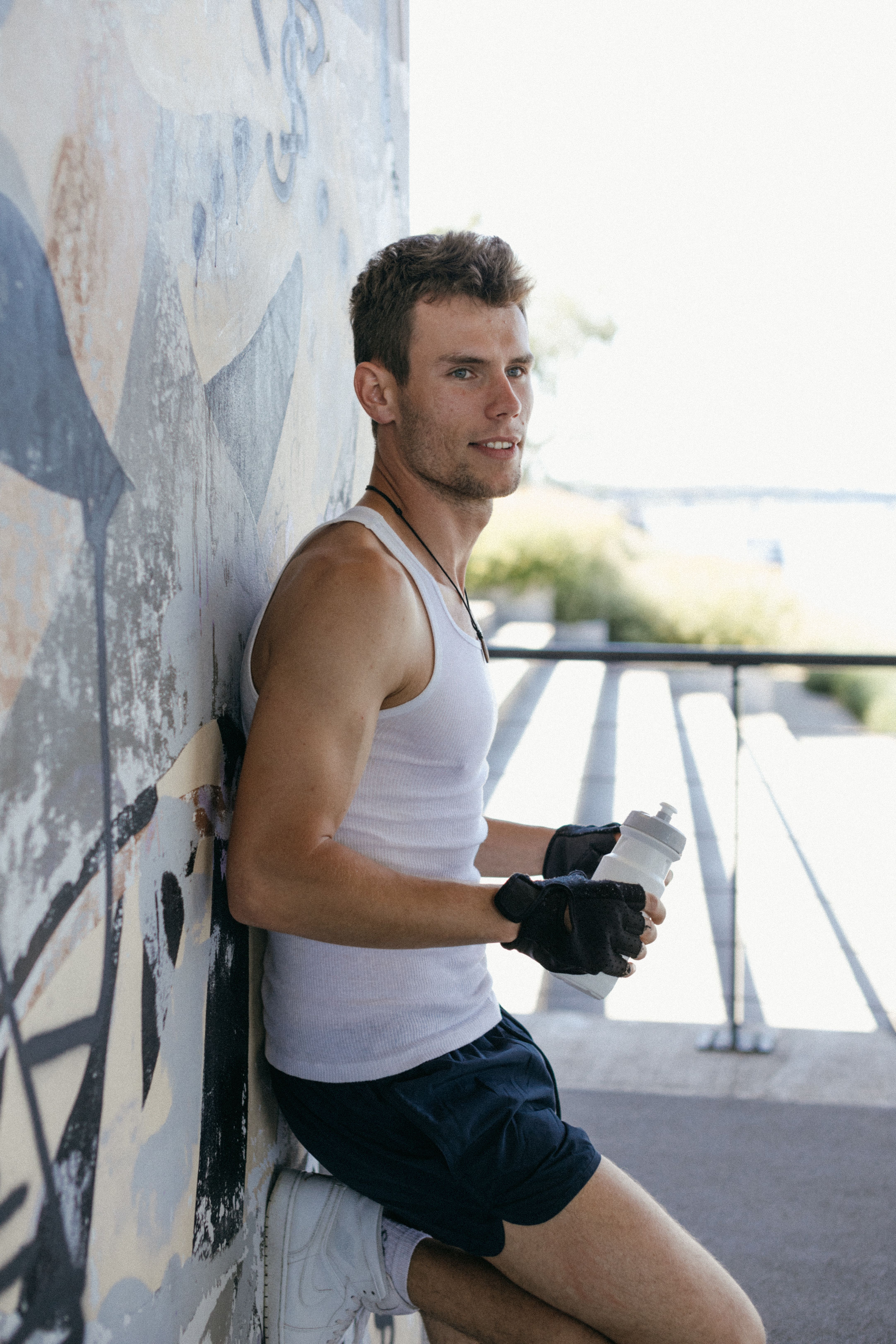 Smiling athletic man in white tank top and shorts holds a water bottle while leaning against a graffiti wall.