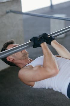Athletic man in tank top exercising with barbell, showcasing fitness and strength indoors.