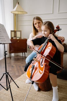 A woman guides a young girl in learning to play the cello indoors.