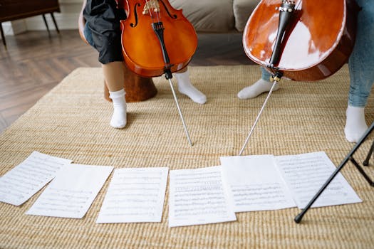 Two people playing cellos with sheet music on the floor during an indoor practice session.