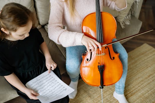 Child in black dress learns cello from teacher in cozy living room setting.
