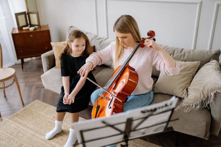 A Woman Playing Cello While Sitting On The Couch Beside The Young Girl In Black Dress