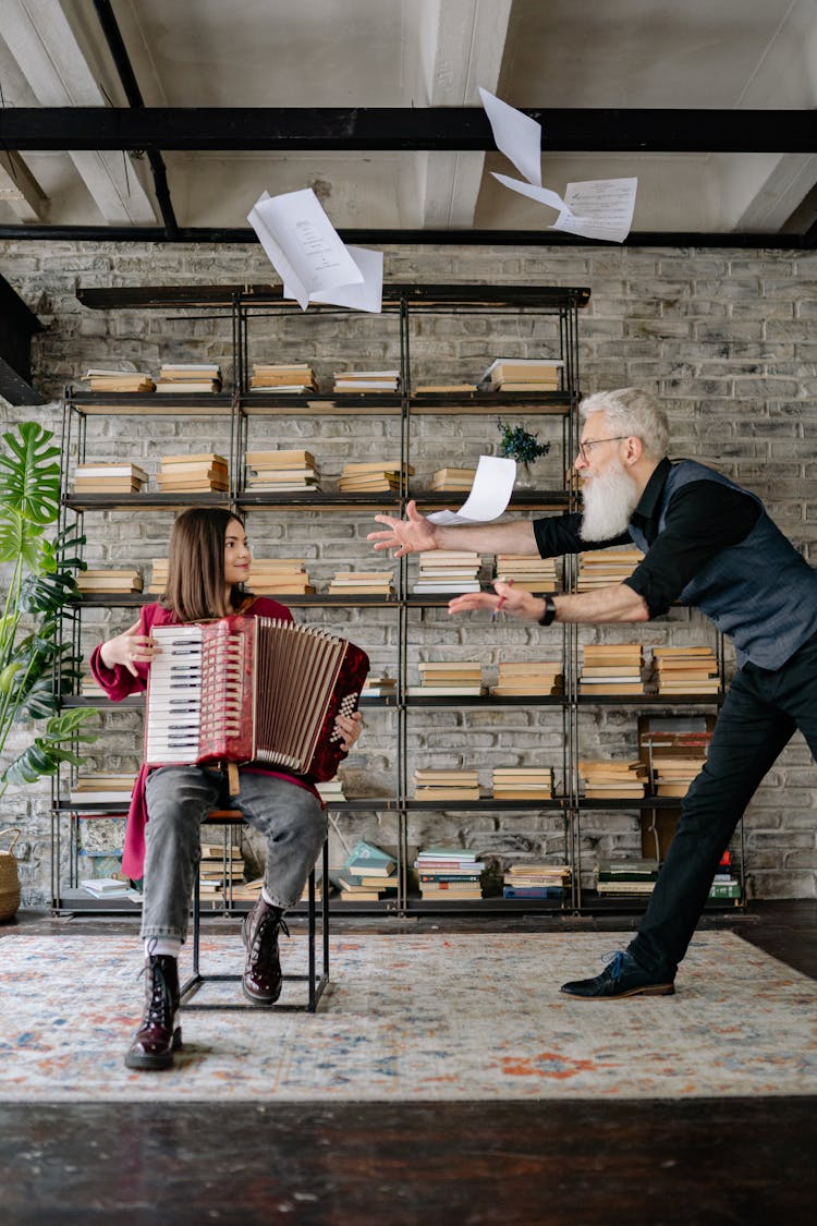 An Elderly Man Throwing Papers To The Girl Sitting On A Chair While Holding Accordion