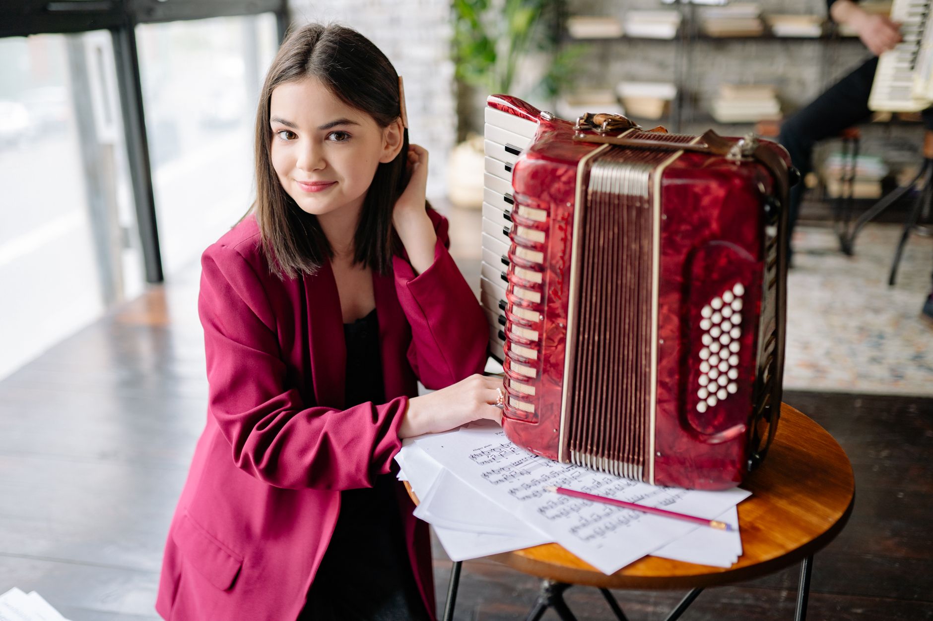 A young woman in a red jacket sits by an accordion, surrounded by music sheets indoors.