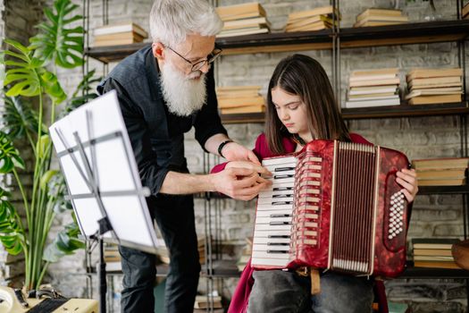 An elderly man teaches a young girl to play the accordion indoors, surrounded by books.