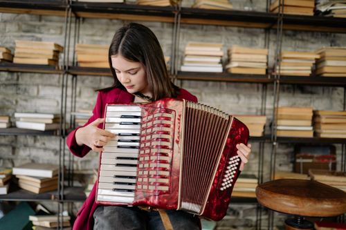 Free A young girl enjoys playing an accordion in a cozy library filled with books, showcasing her musical passion. Stock Photo