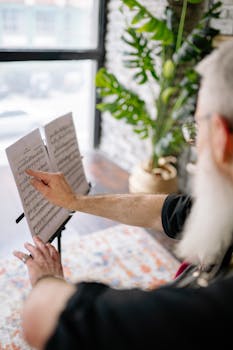 Elderly man with gray beard pointing at sheet music during a music lesson indoors.