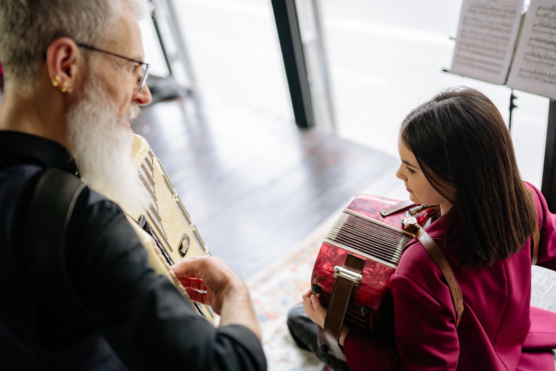 A man teaches a young girl to play the accordion indoors with sheet music nearby.
