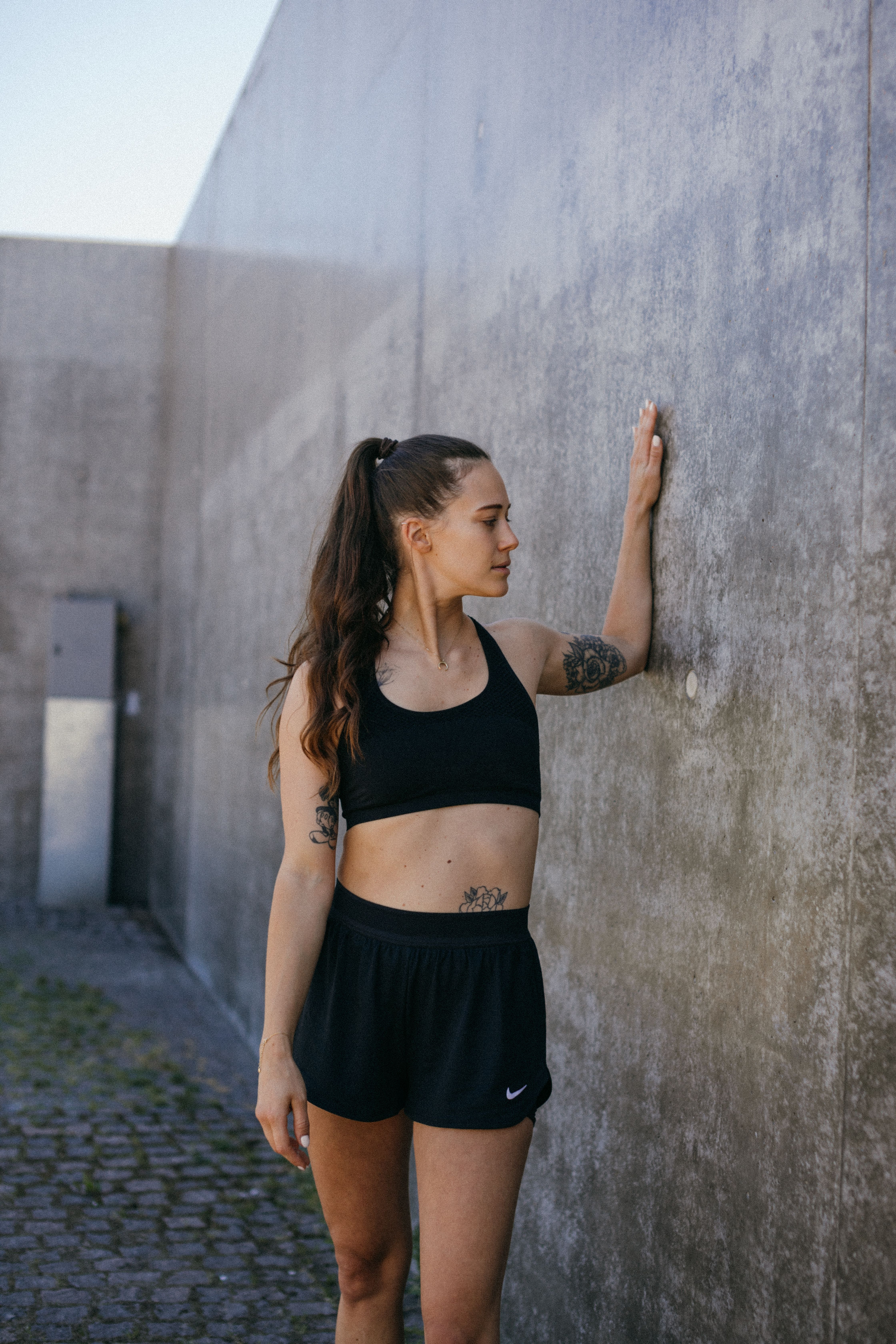 Fit woman in sportswear with tattoos leaning on a concrete wall outdoors.