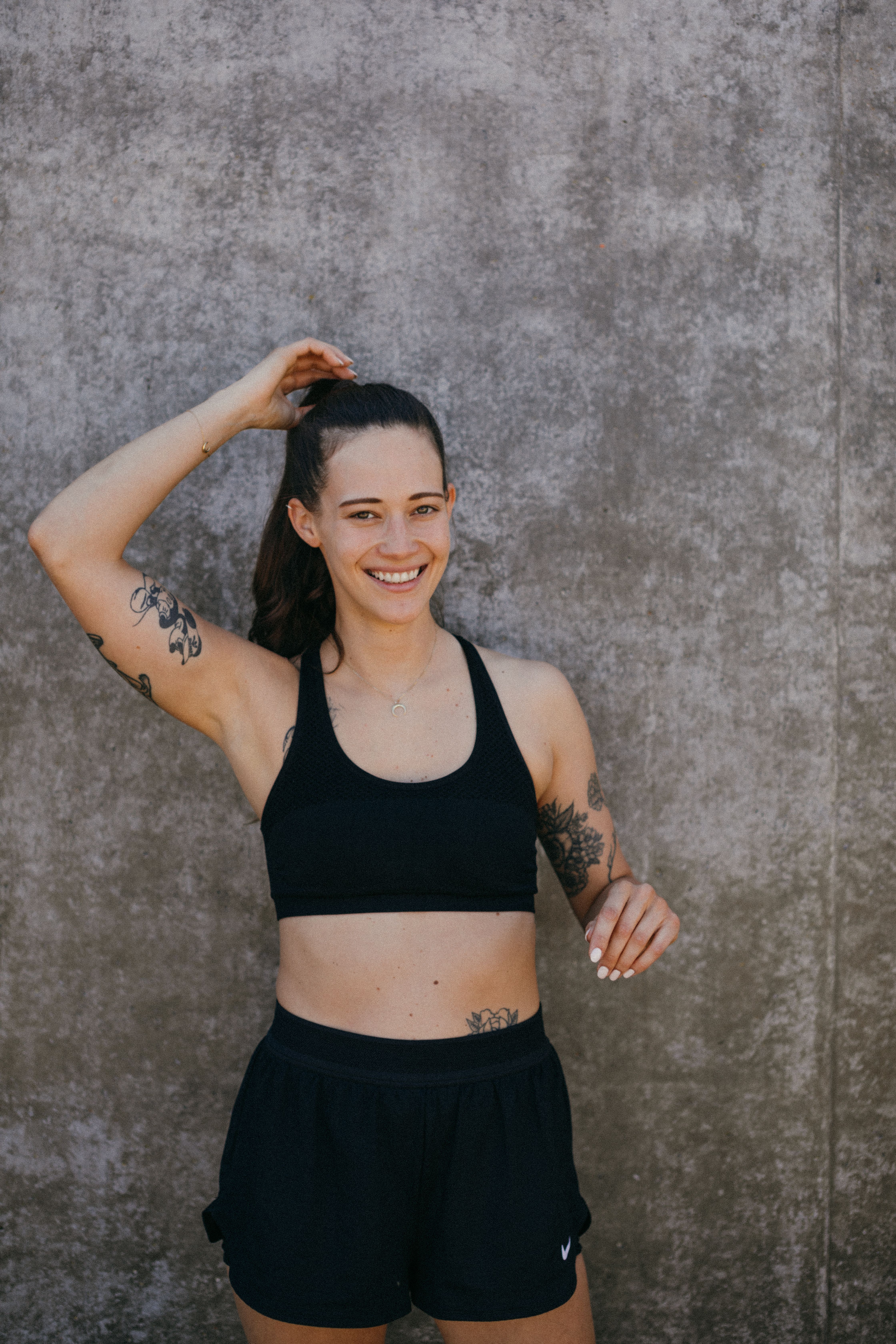 Young woman in black active wear smiling outdoors against concrete wall.