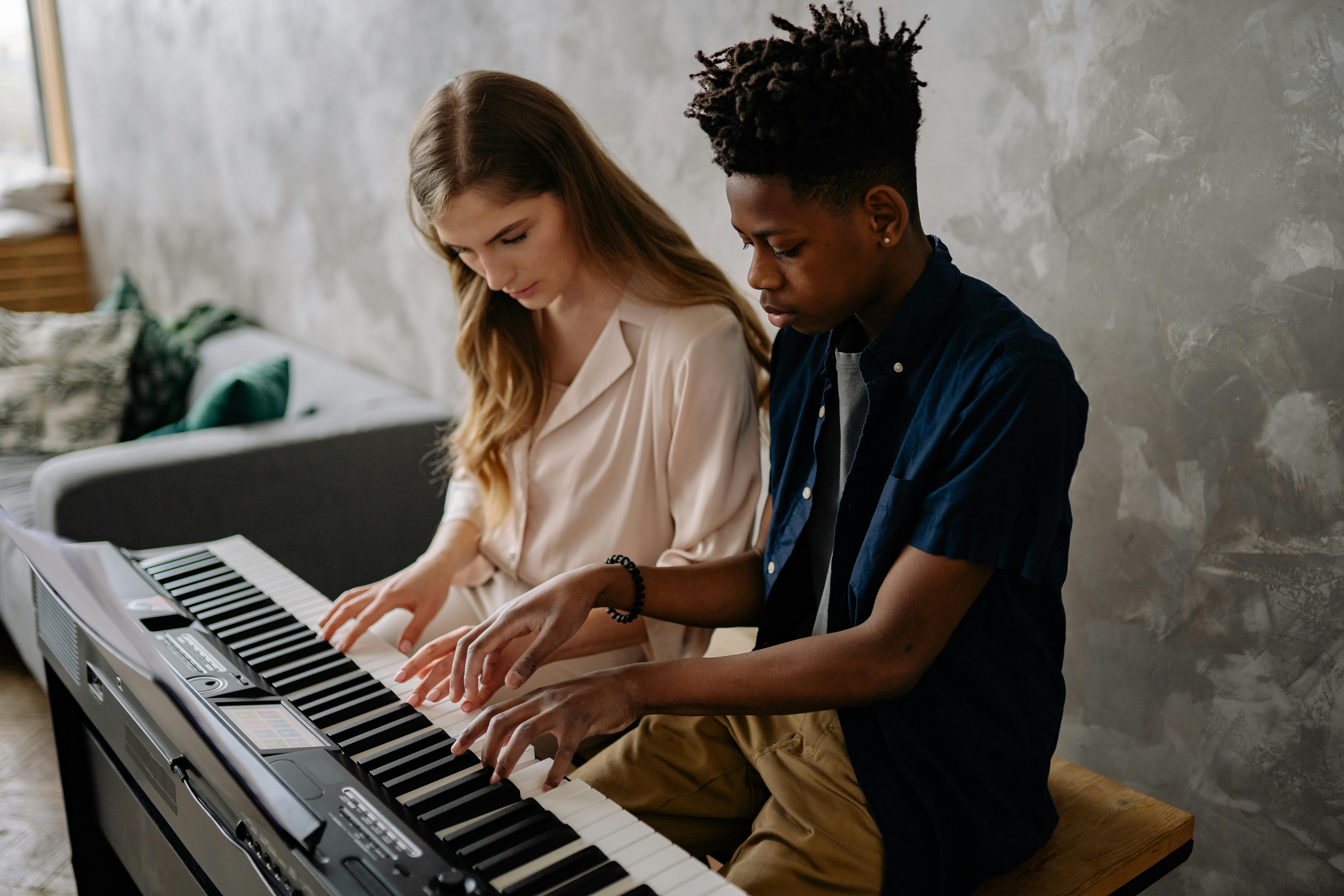 An African American boy and Caucasian woman sit together, playing an electronic keyboard indoors.