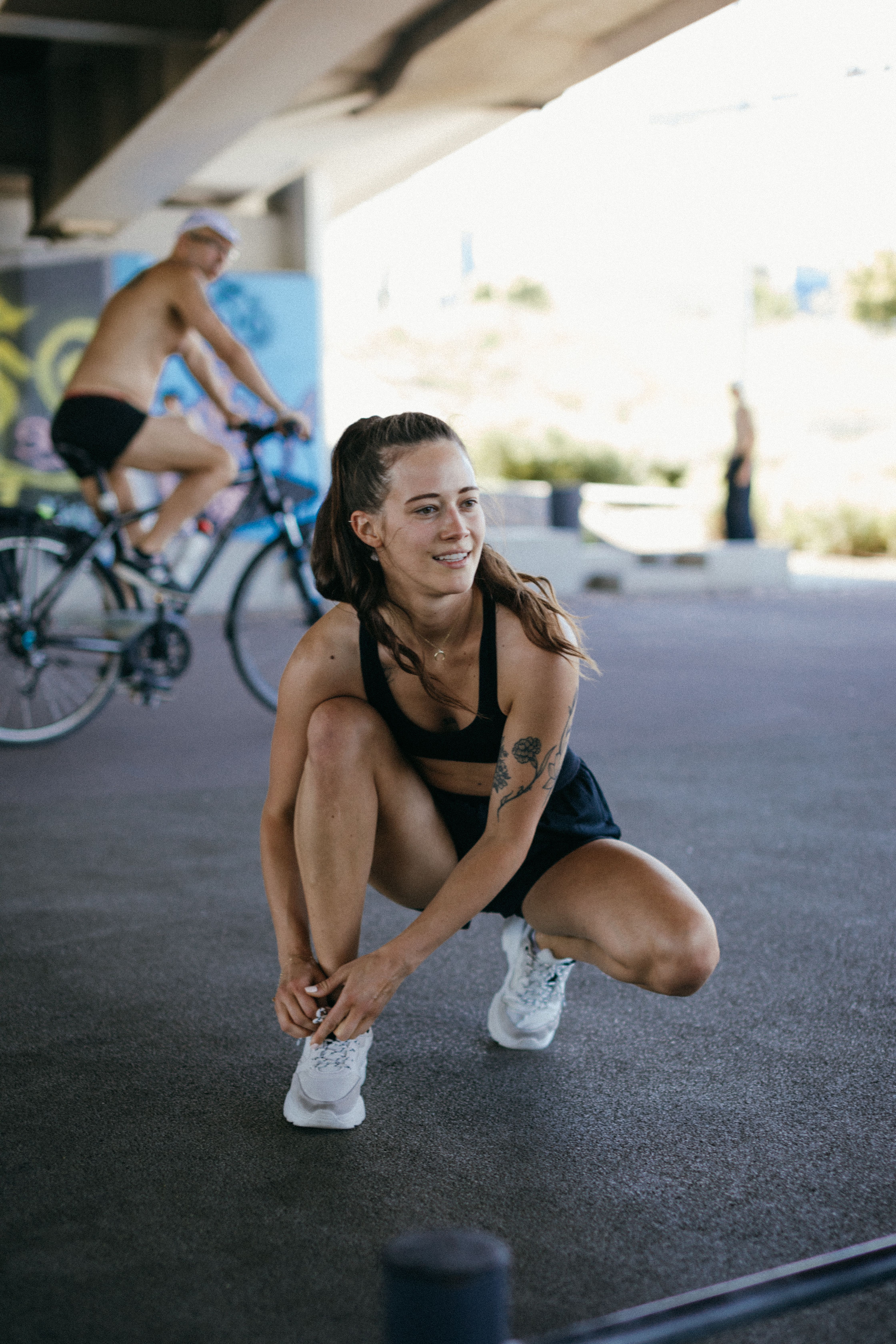 Smiling woman in active wear tying her laces outdoors under a bridge.