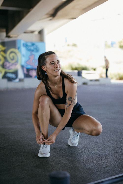 Free Smiling athletic woman ties her shoe under a graffiti-covered bridge, ready for a workout. Stock Photo
