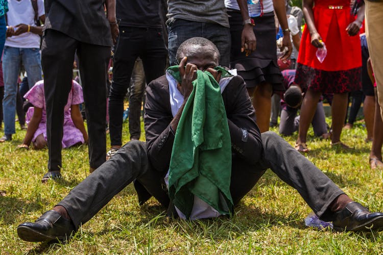 Man Sitting On A Ground With Legs Apart Covering His Face And Crowd In Background