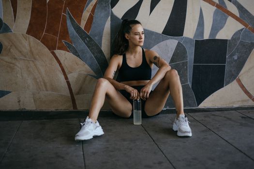 Fitness woman in workout gear resting against a colorful mural with a water bottle.