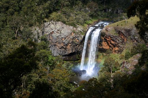 Free A stunning waterfall cascades from rocky cliffs into lush green forest, creating a serene natural scene. Stock Photo