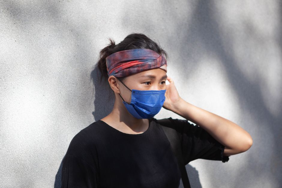 Woman wearing a blue facemask and colorful headband stands by a sunlit wall.