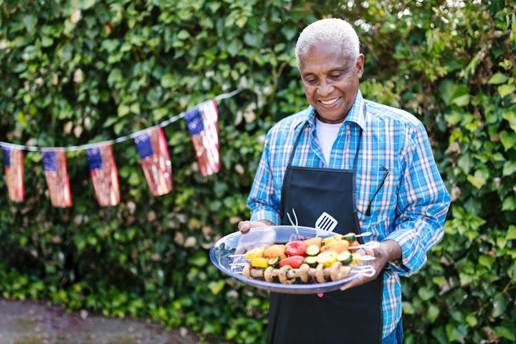 Elderly Man Holding A Tray With Food And Standing In The Garden Decorated For The 4th Of July